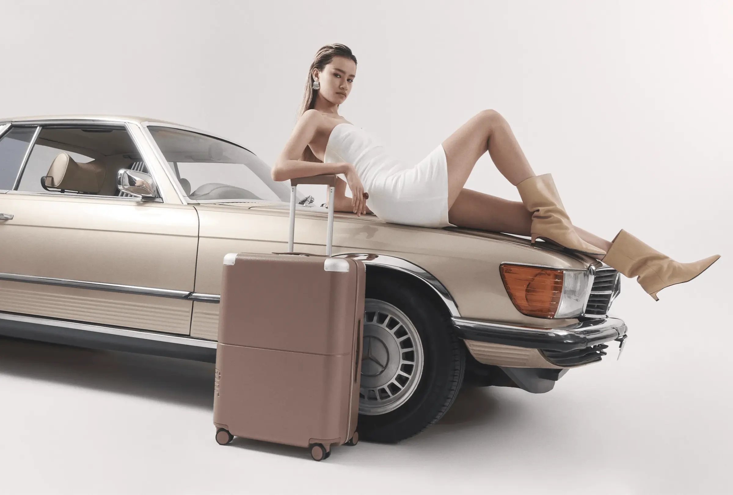 July luggage campaign image with model in white dress reclining on vintage car beside rolling suitcase on minimalist studio background